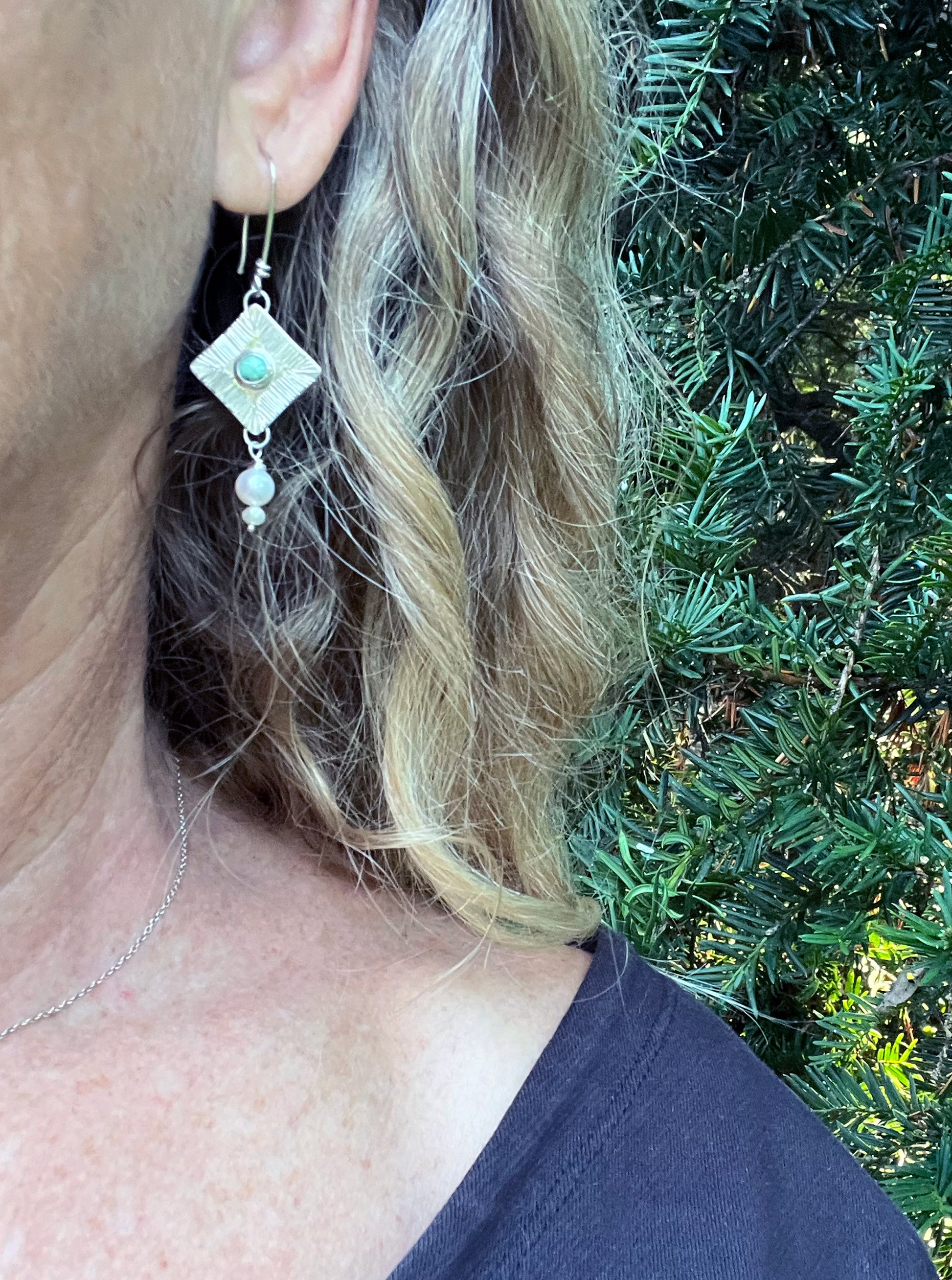 Close-up of a person wearing a silver earring with a variscite stone against a green foliage background